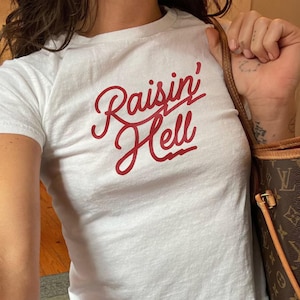 May include: White t-shirt with the red text "Raisin' Hell". The shirt is short-sleeved and appears to be made of cotton. The model is wearing blue jeans and carrying a brown handbag.