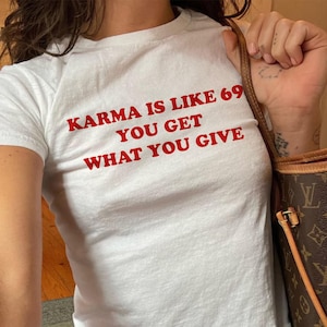 May include: A white short-sleeved t-shirt featuring red text printed on the chest that reads, KARMA IS LIKE 69 YOU GET WHAT YOU GIVE. The shirt is paired with blue denim jeans and a brown patterned shoulder bag with gold-toned hardware accents.
