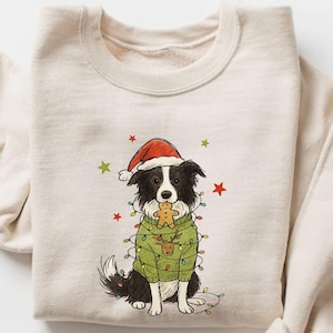Könnte beinhalten: Cremefarbenes Sweatshirt mit einem schwarz-weißen Hund, der einen Weihnachtsmannhut und einen grünen Pullover mit Lichterketten trägt. Der Hund hält einen Lebkuchenmann-Keks. Rote und grüne Sterne sind verstreut.
