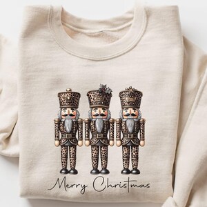 Può includere: Una felpa color crema con tre figure di schiaccianoci con dettagli animalier. Gli schiaccianoci hanno dettagli dorati e sono allineati. Le parole "Merry Christmas" sono scritte sotto in un carattere corsivo.