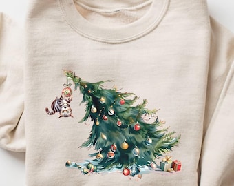 May include: A cream-colored sweatshirt featuring a festive Christmas tree with ornaments and a playful cat. The graphic includes wrapped gifts at the base of the tree, creating a cozy holiday theme.