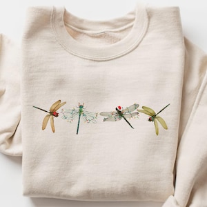 Può includere: Felpa color crema con girocollo e maniche lunghe. Presenta un ricamo di quattro libellule nei toni del marrone, verde e grigio, con dettagli rossi su alcune.