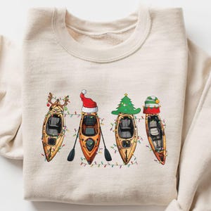 以下が含まれることがあります： ベージュ色のスウェットシャツ。クリスマスデコレーションで飾られた4つのカヤックの、お祭りデザインが特徴です。各カヤックには、トナカイの角、サンタの帽子、クリスマスツリー、ストライプの帽子など、ユニークなホリデーテーマがあります。カヤックはクリスマスライトで飾られています。