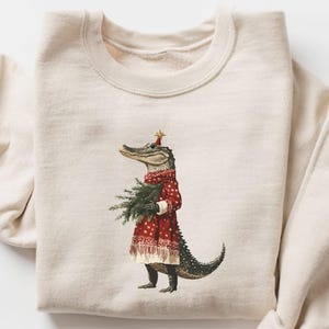 Op de afbeelding: Crèmekleurige sweatshirt met een grillige illustratie van een alligator gekleed in een rood-wit patroonjas en een met sterren bekroonde hoed, met een bundel dennentakken. Het ontwerp heeft een feestelijk vakantiethema.