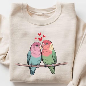 May include: Beige sweatshirt featuring a colorful illustration of two lovebirds perched on a branch, with hearts above them. The birds have pink, blue, and green feathers. The sweatshirt has long sleeves and a crew neck.