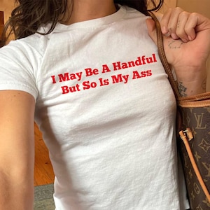 May include: White t-shirt with the text "I May Be A Handful But So Is My Ass" in red. The shirt is paired with blue jeans. A brown handbag with a monogram pattern is visible.