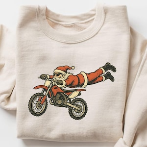 Puede incluir: Sudadera color crema con un diseño de Papá Noel montando una moto de cross roja. Papá Noel lleva gafas de sol y un traje rojo. El diseño tiene un estilo vintage de dibujos animados, ideal para Navidad o festividades.