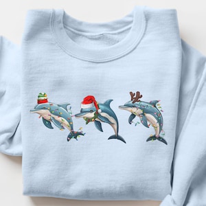 Peut inclure: Sweat-shirt bleu clair orné de trois dauphins de dessin animé décorés pour Noël. Chaque dauphin porte un chapeau ou des bois de renne festifs et est orné de guirlandes lumineuses. Le motif est centré sur le devant.