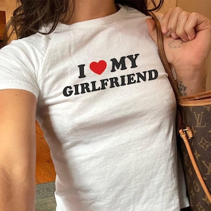 May include: White t-shirt with the text "I ❤️ MY GIRLFRIEND" in black letters and a red heart. The shirt is paired with blue jeans. A brown handbag with a monogram pattern is visible.