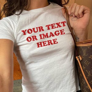 May include: White t-shirt with the text "YOUR TEXT OR IMAGE HERE" in red. The shirt is a crew neck style and is paired with blue jeans. A brown handbag with a monogram pattern is visible.