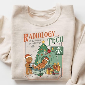 Puede incluir: Sudadera color crema con un gráfico festivo. El diseño presenta figuras de pan de jengibre con gorros de Papá Noel, un árbol de Navidad y una máquina de resonancia magnética. El texto dice "Radiology Tech" y "'Tis the season to radiate joy."