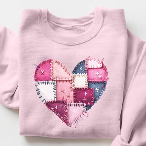 Peut inclure: Sweat-shirt rose clair orné d'un cœur en patchwork. Le cœur est composé de carrés de tissu roses, bleus et blancs, cousus ensemble avec du fil visible. Le texte "LIGHT PINK" est en bas.