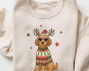 May include: Cream-colored sweatshirt featuring a cartoon dog wearing a Christmas sweater, heart-shaped sunglasses, and reindeer antlers with lights. The dog is surrounded by red and green stars. A festive holiday design.