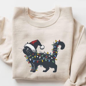 以下が含まれることがあります： クリーム色のスウェットシャツ。サンタ帽をかぶり、カラフルなクリスマスライトで飾られた黒猫のイラストが描かれています。猫は漫画風で、丸まった尻尾とひげがあります。スウェットシャツはクルーネックと長袖です。