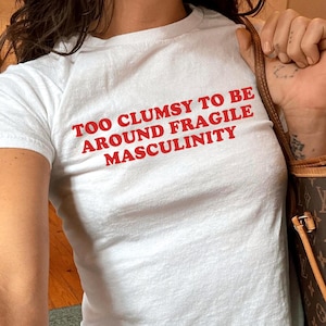 May include: White t-shirt with the red text "TOO CLUMSY TO BE AROUND FRAGILE MASCULINITY." The shirt is a crew neck style. The model is wearing blue jeans. A brown and tan handbag is in the background.