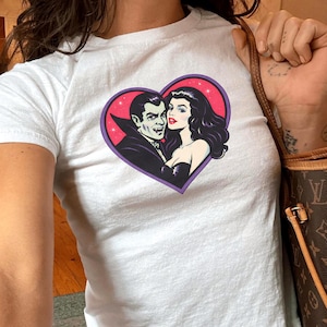 Könnte beinhalten: Weißes T-Shirt mit einem herzförmigen Aufdruck eines Vampirs und einer Frau. Die Grafik ist lila und rosa umrandet. Das T-Shirt wird mit einer blauen Jeans kombiniert. Das Model trägt Größe XS.