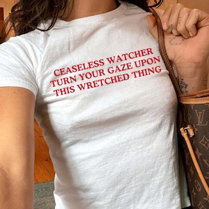 Op de afbeelding: Wit T-shirt met de rode tekst "CEASELESS WATCHER TURN YOUR GAZE UPON THIS WRETCHED THING." Het shirt wordt gecombineerd met een blauwe jeans. Een bruine en beige handtas is zichtbaar.
