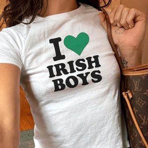 Może przedstawiać: Biała koszulka z napisem "I ♥ IRISH BOYS" czarnymi literami i zielonym sercem. Koszulka jest połączona z niebieskimi dżinsami. Po prawej stronie zdjęcia widoczna jest brązowa torebka.