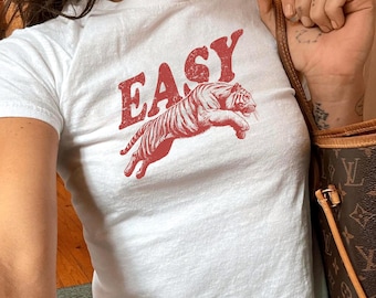May include: White t-shirt with a red graphic of a tiger leaping and the word "EASY" above it. The shirt has short sleeves and a crew neck. The model is wearing blue jeans.