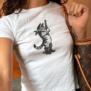 Könnte beinhalten: Weißes kurzärmeliges T-Shirt mit einer schwarz-weißen Grafik einer Katze, die eine Pistole hält, ein Halstuch und Cowboy-Stiefel trägt. Die Katzen-Grafik ist mittig auf der Vorderseite des Shirts platziert. Das Shirt wird mit einer blauen Jeans kombiniert.