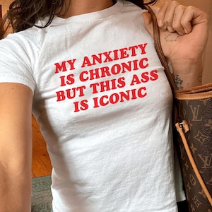 Puede incluir: Camiseta blanca con el texto rojo "MY ANXIETY IS CHRONIC BUT THIS ASS IS ICONIC." La camiseta se combina con vaqueros azules. La modelo lleva la talla XS y pesa unos 54 kg.