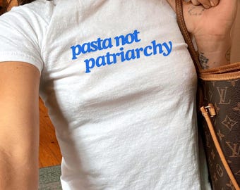 Pode incluir: Camiseta branca com o texto azul "pasta not patriarchy". A camisa é de manga curta e parece ser feita de um material macio. A modelo está usando jeans azuis e carregando uma bolsa marrom.