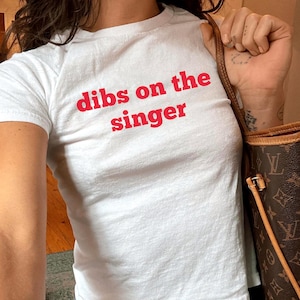 May include: White t-shirt with the phrase "dibs on the singer" in red lettering. The shirt is paired with blue jeans. A brown and tan handbag is visible in the image.