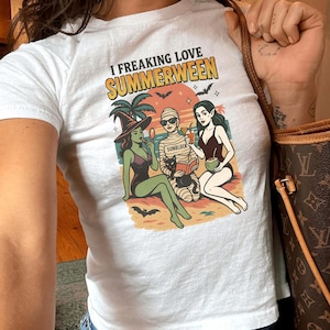 Op de afbeelding: Wit T-shirt met de tekst "I FREAKING LOVE SUMMERWEEN" en een afbeelding van drie stripfiguren in badkleding. Eén is een heks, één is een mummie en één is een vrouw. De mummie heeft het woord "SUNBLOCK" op zijn verband.