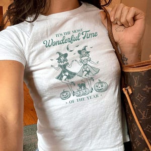 Puede incluir: Camiseta blanca con un gráfico verde azulado que dice "IT'S THE MOST Wonderful Time + OF THE YEAR +." El gráfico presenta dos figuras de brujas bailando, calabazas y una luna creciente. La camiseta se combina con vaqueros azules.