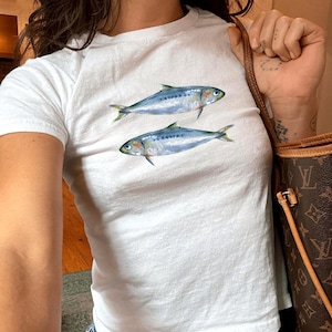 May include: White t-shirt featuring two watercolor-style fish. The fish are blue and silver with yellow fins. The shirt is paired with blue jeans. The model is wearing size XS.
