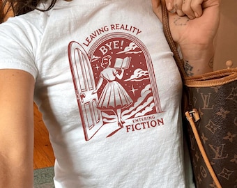 May include: White t-shirt featuring a red graphic of a woman entering a door, holding a book. The text on the shirt says "LEAVING REALITY BYE! ENTERING FICTION". The shirt is worn with blue jeans.