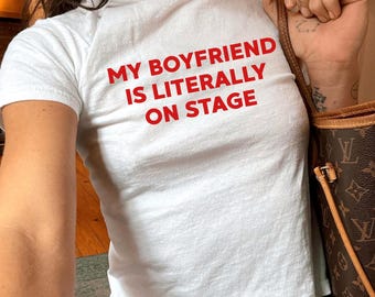 Puede incluir: Camiseta blanca con el texto rojo "MY BOYFRIEND IS LITERALLY ON STAGE." La camiseta se combina con unos vaqueros azules. Se ve un bolso marrón con una correa marrón claro.