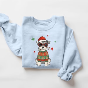 May include: Light blue sweatshirt featuring a cartoon Schnauzer wearing a Santa hat, sunglasses, and a Christmas sweater with gingerbread men. The design includes red and green stars, and a candy cane.