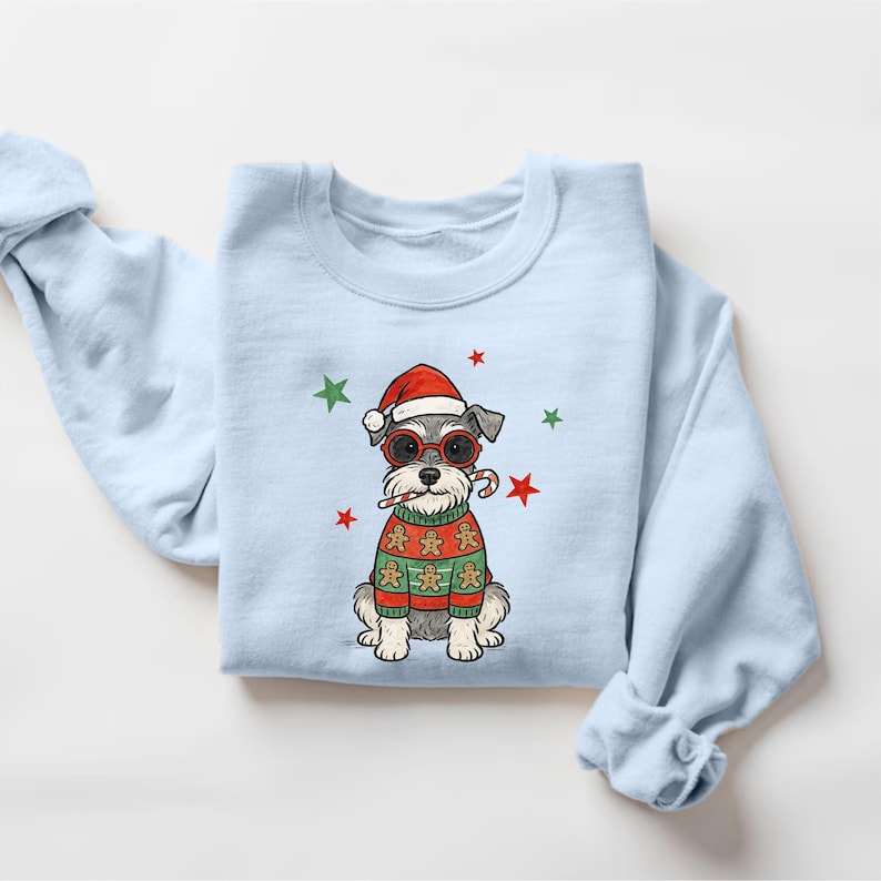 May include: Light blue sweatshirt featuring a cartoon Schnauzer wearing a Santa hat, sunglasses, and a Christmas sweater with gingerbread men. The design includes red and green stars, and a candy cane.