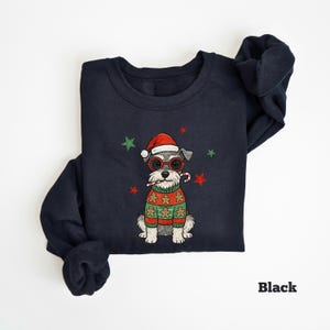 May include: Black sweatshirt featuring a Schnauzer dog wearing a Santa hat, red glasses, and a gingerbread sweater. The dog is holding a candy cane. Green and red stars are scattered around the dog. The word "Black" is printed at the bottom right.