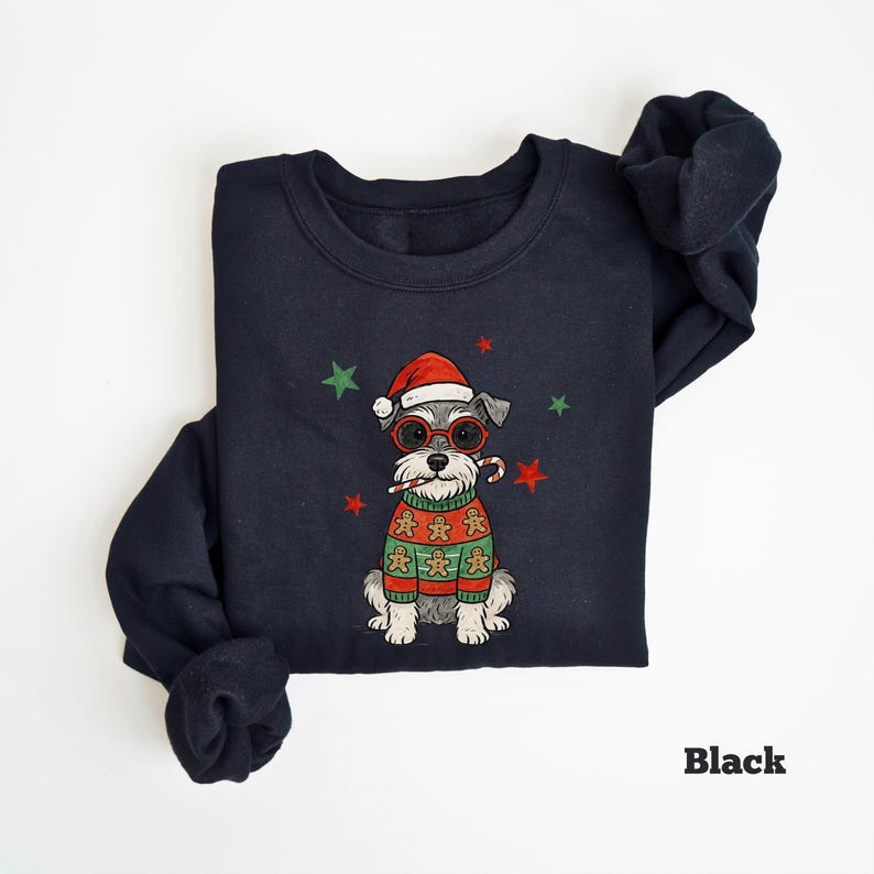 May include: Black sweatshirt featuring a Schnauzer dog wearing a Santa hat, red glasses, and a gingerbread sweater. The dog is holding a candy cane. Green and red stars are scattered around the dog. The word "Black" is printed at the bottom right.