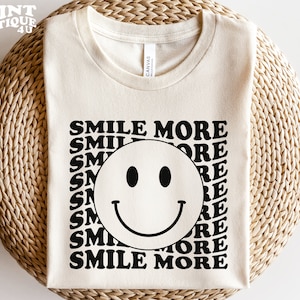 Puede incluir: Camiseta color crema con las palabras "SMILE MORE" en un patrón repetido alrededor de una gran cara sonriente. La camiseta está hecha de un material suave y tiene un cuello redondo clásico.