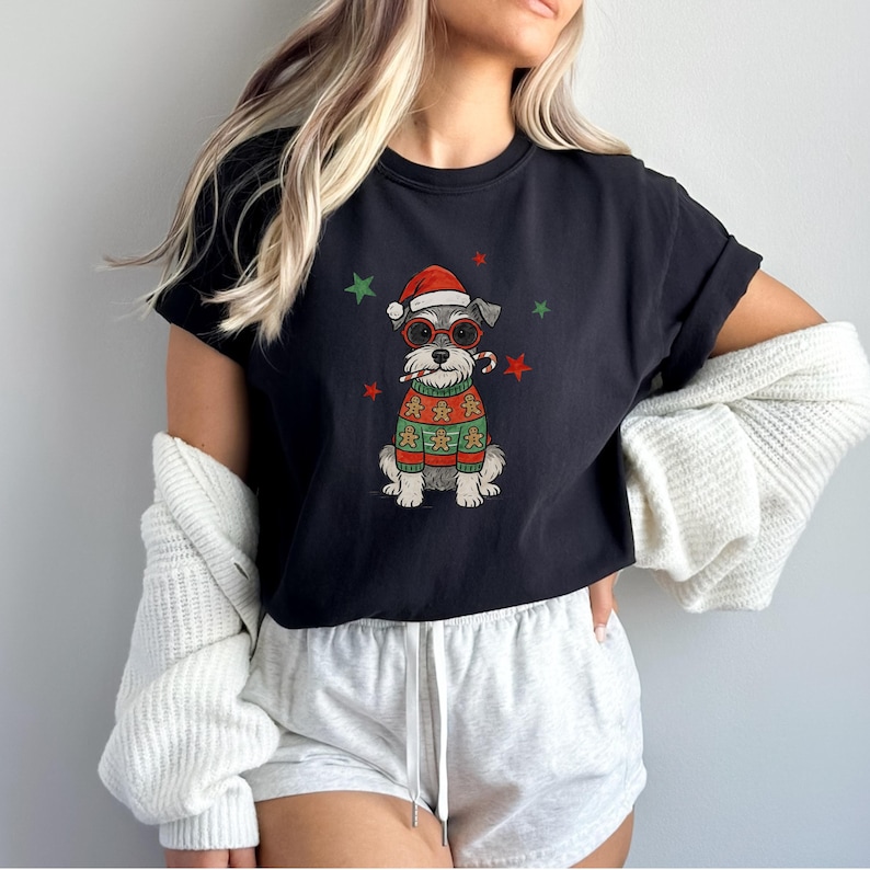 May include: Black t-shirt featuring a Schnauzer dog wearing a Santa hat, red glasses, and a gingerbread sweater. The dog is holding a candy cane. Green and red stars are scattered around the dog. A festive holiday design.