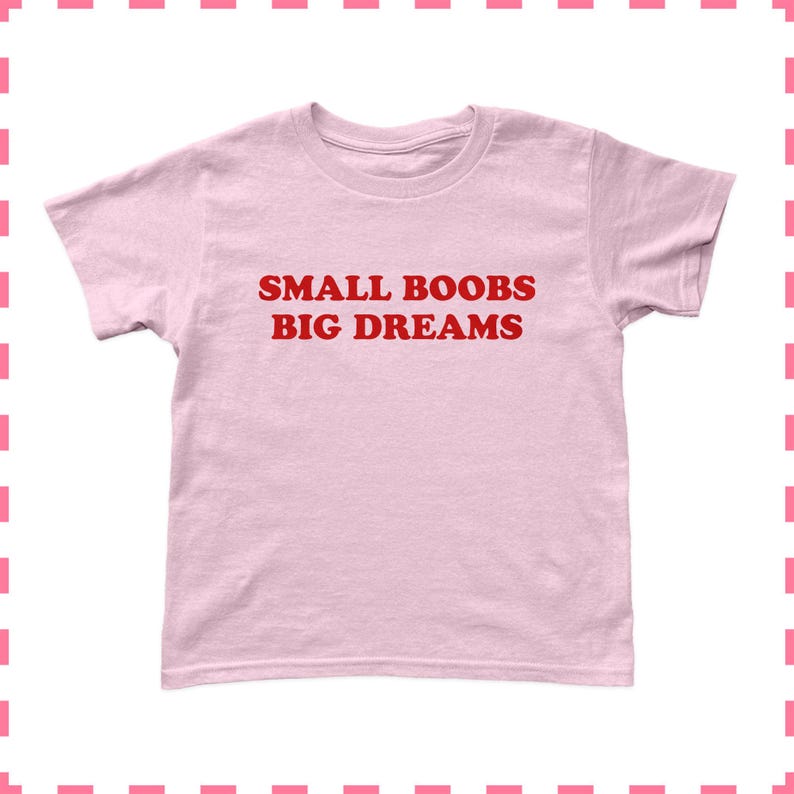 May include: A light pink t-shirt with the text "SMALL BOOBS BIG DREAMS" in red block letters. The shirt is a crew neck style and appears to be made of a soft material. The shirt is a solid color.