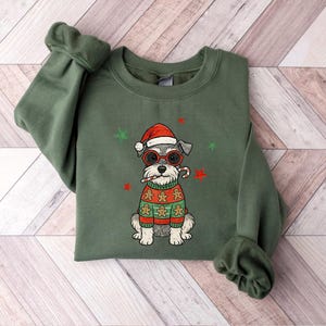 May include: An olive green sweatshirt featuring a Schnauzer dog wearing a Santa hat, red glasses, and a gingerbread sweater. The dog is holding a candy cane. Green and red star accents are scattered around the dog.