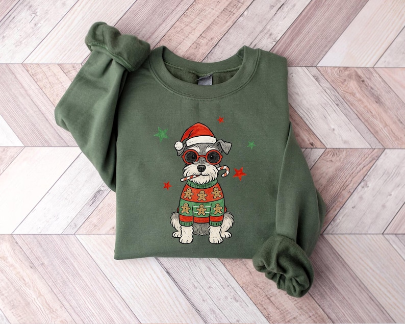 May include: An olive green sweatshirt featuring a Schnauzer dog wearing a Santa hat, red glasses, and a gingerbread sweater. The dog is holding a candy cane. Green and red star accents are scattered around the dog.