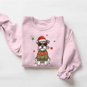 May include: A light pink sweatshirt featuring a cartoon dog wearing a Santa hat, sunglasses, and a gingerbread sweater. The dog is holding a candy cane. Green and red stars decorate the background. A festive holiday design.