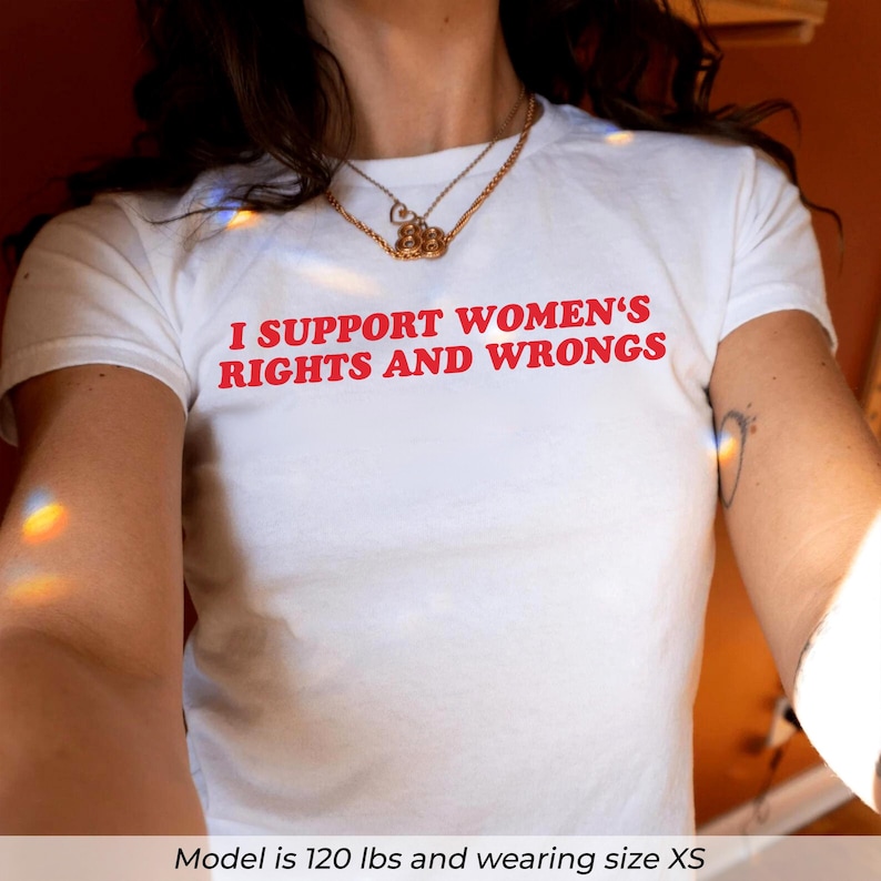 May include: White t-shirt with the red text "I SUPPORT WOMEN'S RIGHTS AND WRONGS". The shirt is a crew neck style. A gold necklace is worn around the neck.
