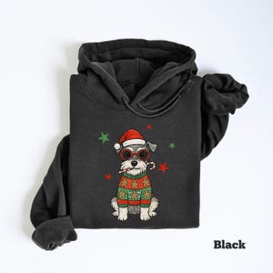May include: Black hoodie featuring a cartoon dog wearing a Santa hat, glasses, and a gingerbread sweater. The dog is holding a candy cane. Green and red stars are scattered around the dog. The word "Black" is printed at the bottom.