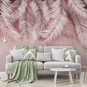 Pu&ograve; includere: Un divano grigio con una coperta verde e cuscini con motivi geometrici e a foglia si trova di fronte a un muro con un design a foglie di palma rosa e bianco.