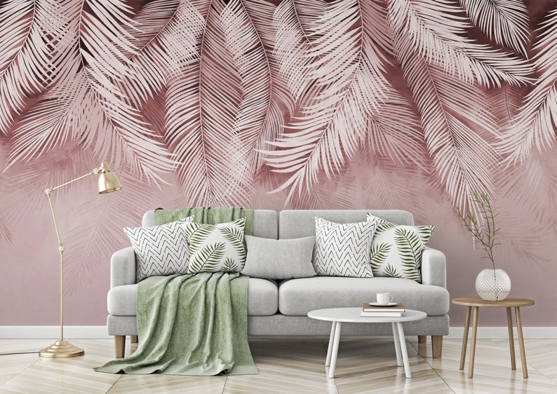 Pu&ograve; includere: Un divano grigio con una coperta verde e cuscini con motivi geometrici e a foglia si trova di fronte a un muro con un design a foglie di palma rosa e bianco.