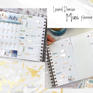 May include: A mini planner with a blue and white cover featuring a beach scene. The planner has a July calendar page and a blank page with a dotted grid. The planner is decorated with stickers and washi tape.
