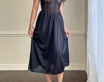 Puede incluir: Un vestido de encaje y satén negro con escote en V y falda fruncida. El vestido es sin mangas y tiene un corpiño ajustado. La modelo lleva tacones negros.