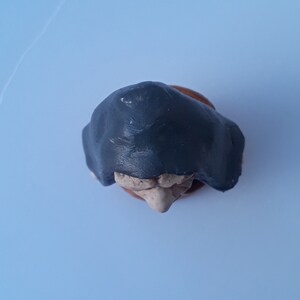 May include: A small ceramic figurine of a person wearing a dark gray hooded cloak. The figurine is made of clay and has a textured surface.