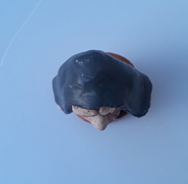 May include: A small ceramic figurine of a person wearing a dark gray hooded cloak. The figurine is made of clay and has a textured surface.